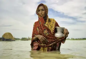 A woman walks in water carrying drinking water, in Bangladesh
